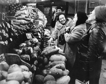 Black and white photograph showing four figures laughing, looking at produce in a market stall.