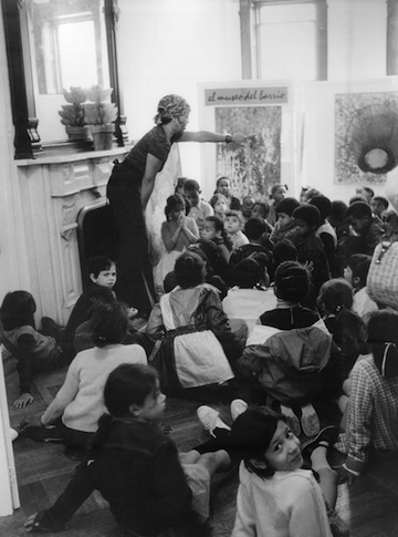 Black and white photograph showing a room of children sitting on the floor with an adult figure standing in the middle, pointing at a pair of artworks.