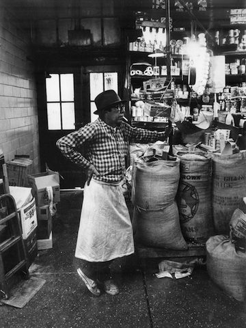 Black and white photograph of a market vendor standing in front of bags of rice.