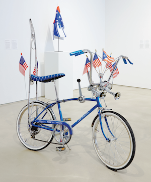 Photograph of a bicycle with decked with flags in the forefront, and a Klansman hood in the background.