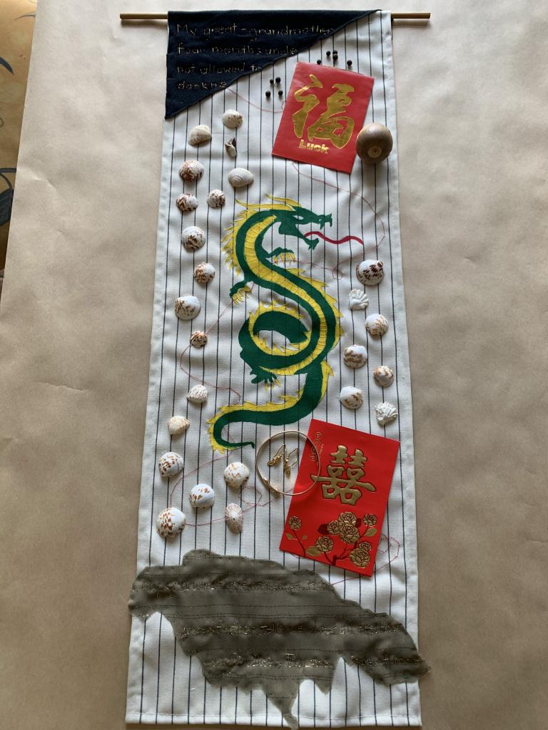 "mixed media artwork of cloth, thread, shells, red envelopes and seeds with green and yellow painted dragon in center"